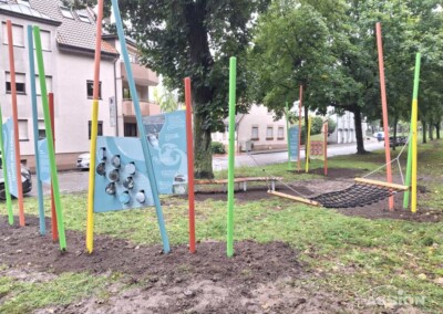 Spielplatz im Freien mit bunten vertikalen Stangen, einer Kletterwand, einer Hängematte aus Seilnetzen und Informationsschildern auf einer Wiese in der Nähe von Bäumen und einem Wohngebäude.