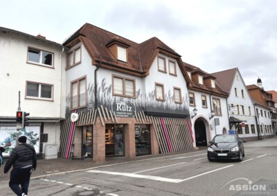 Eine Bäckerei mit dem Schriftzug "Rütz" befindet sich im Erdgeschoss eines Eckhauses an einer Stadtstraße; ein Auto fährt vorbei und ein Fußgänger überquert die Straße.
