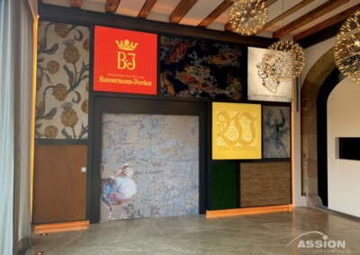 Wand mit verschiedenen dekorativen Paneelen, darunter beleuchtete Logos der Weinkellerei, florale Muster, ein abstraktes Blau-Weiß-Motiv und Holzstrukturen; von der Decke hängen moderne Beleuchtungsarmaturen.