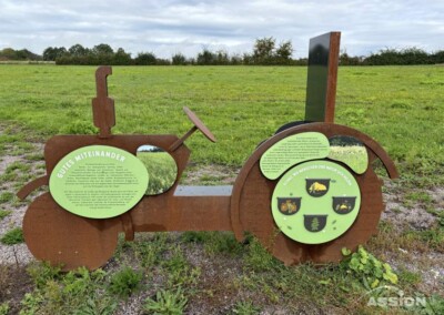 Eine rostfarbene Traktorskulptur aus Metall mit grünen Hinweisschildern steht auf einem Kiesplatz in einem grasbewachsenen Feld unter einem bewölkten Himmel.
