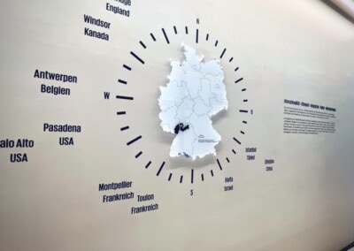 Wanddisplay mit einer Deutschlandkarte, umgeben von Städtenamen aus verschiedenen Ländern, darunter England, Kanada, Belgien, USA und Frankreich, verbunden durch radiale Linien.