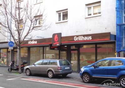 Straßenansicht des Restaurants Grillhaus Medina mit geparkten Autos, einem Motorrad und einem Fahrradparkschild vor einem beigen Gebäude.