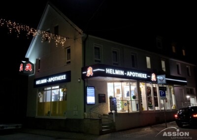 Eine Apotheke namens Helmin-Apotheke ist nachts mit sichtbaren Schildern und Innenleuchten beleuchtet; einige Weihnachtslichter hängen über der Straße.