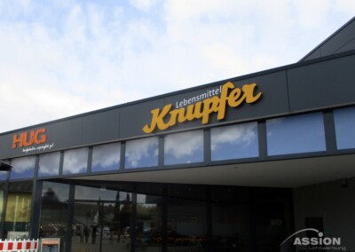 Ein modernes Schaufenster mit den Schildern "HUG" und "Lebensmittel Knupfer" unter einem wolkenlosen blauen Himmel.