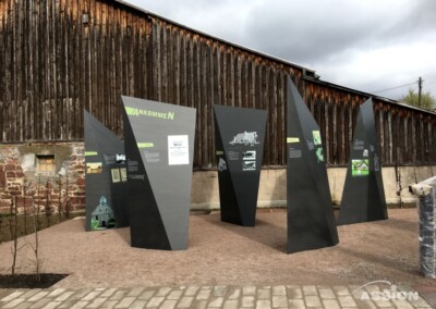 Freiluftausstellung mit mehreren hohen, schwarzen, kantigen Informationstafeln vor einem verwitterten Holzgebäude, auf einem Kiesplatz mit einem gepflasterten Weg im Vordergrund.