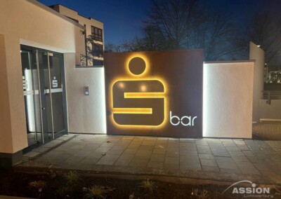 Beleuchtetes Schild mit einem großen stilisierten "S" und dem Wort "Bar" an einer Wand außerhalb eines Gebäudes bei Nacht. In der Nähe befindliche Glastüren zeigen das gleiche "S"-Symbol.