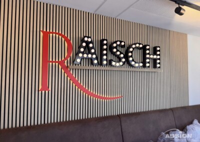 Wandschild mit großem rotem "R" und Leuchtbuchstaben mit der Aufschrift "AISCH" auf einem vertikalen Lamellenhintergrund über einer braunen gepolsterten Bank.