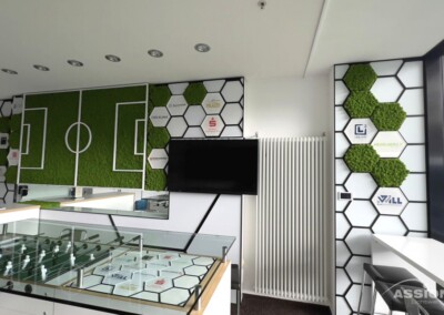 Moderner Büroraum mit sechseckigen Wandpaneelen, grünen Moos-Akzenten, einem montierten Fernseher, einem Kickertisch und einer Glasvitrine.