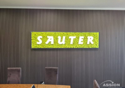 Ein Schild mit dem Wort "SAUTER" in weißen Buchstaben auf grünem Grund ist an einer Wand über drei braunen Stühlen und einem Tisch angebracht.