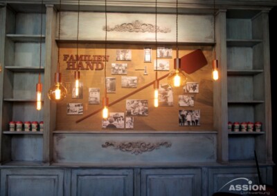 Ein Wanddisplay mit alten Fotos, einem großen Holzpaddel und hängenden Edison-Glühbirnen, umgeben von Regalen mit kleinen Gläsern mit roten Deckeln.