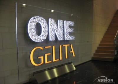 Beleuchtetes Wandschild mit der Aufschrift "ONE GELITA" mit "ONE" in Weiß und "GELITA" in Orange, neben einer Treppe in einem modernen Gebäude.