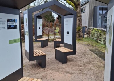 Außeninstallation mit modernen, unterstandsähnlichen Strukturen, Holzbänken und Informationstafeln, die auf einem Kiesweg in der Nähe von Bäumen und einem Gebäude stehen.