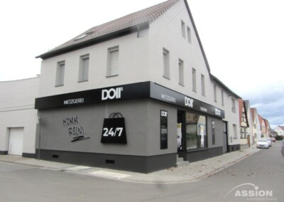 Ein Eckgebäude mit einer Metzgerei namens "DOIT Metzgerei" mit schwarzer Beschilderung und einer großen "24/7"-Grafik. Die Straße scheint ruhig zu sein, es sind keine Menschen anwesend.