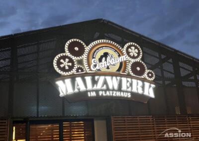 Leuchtreklame für "Eichbaum Malzwerk im Platzhaus" mit zahnradförmigen Verzierungen, montiert auf einer Metallgitterfassade in der Abenddämmerung.