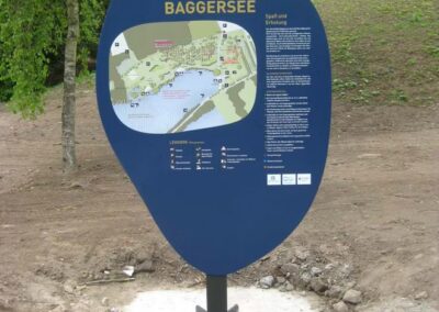 Ein blaues Informationsschild zeigt eine Karte und Details über das Germersheimer Baggerseegebiet an. Es steht auf einem gepflasterten Weg mit Bäumen und Gras im Hintergrund.