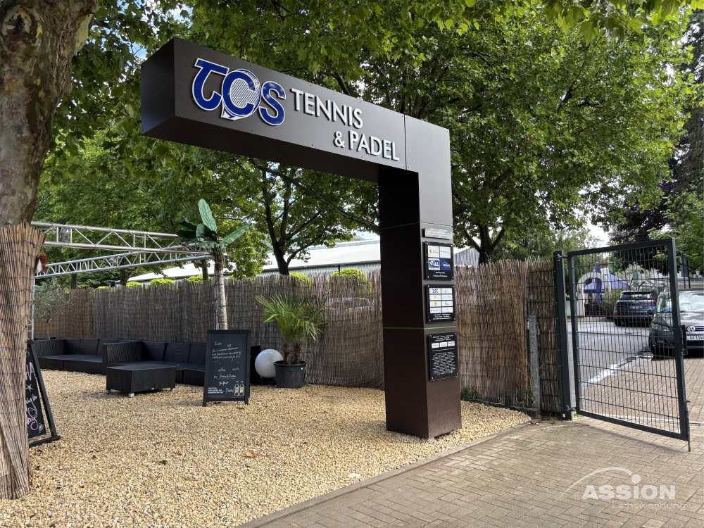 Ständerwerke (1) XXX Eingang zu einem Tennis- und Padel-Club mit schwarzem Schild TCS Tennis & Padel", Sitzgelegenheiten im Freien und einem offenen Tor in einer grünen Umgebung, die durch moderne Trockenbau- und Metallständerwerk-Elemente aufgewertet wird.
