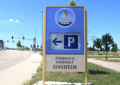 Das Außenschild der Thermen & Badewelt Sinsheim zeigt einen Pfeil nach links und ein Parksymbol, das an einer Straße an einem sonnigen Tag steht.