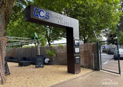 Eingang zu einem Tennis- und Padelclub mit einem großen Schild mit der Aufschrift "TCS Tennis & Padel", Sitzgelegenheiten im Freien und einem eingezäunten Tor.