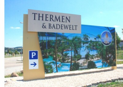 Großes Außenschild für "Thermen & Badewelt" mit einem Parksymbol, das ein Foto eines tropischen Hallenbadkomplexes im Hintergrund zeigt.