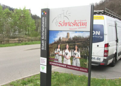Außenschild in Schriesheim, das für die Schriesheimer Weinhoheiten 2024/25 wirbt. Es zeigt drei Frauen in traditioneller Kleidung mit Weinbergen im Hintergrund.