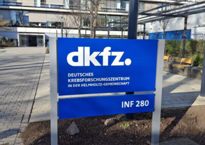 Blaues Schild des Deutschen Krebsforschungszentrums mit der Adresse INF 280 vor einem modernen Bürogebäude.