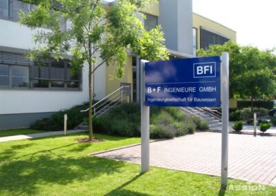 Ein blaues Schild mit der Aufschrift "B+F INGENIEURE GMBH" steht vor einem modernen Bürogebäude mit begrüntem Rasen und Bäumen.