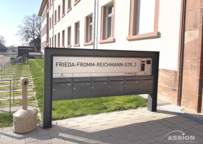 Ein Schild mit Briefkästen zeigt an einem sonnigen Tag die Adresse "Frieda-Fromm-Reichmann-Str. 2" vor einem mehrstöckigen Gebäude an.