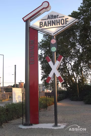 Ein Schild mit der Aufschrift "Alter BAHNHOF" und dem Symbol eines Bahnübergangs steht neben einer roten vertikalen Markierung mit der Aufschrift "Eichbaum" an einem Schotterweg und Bäumen.