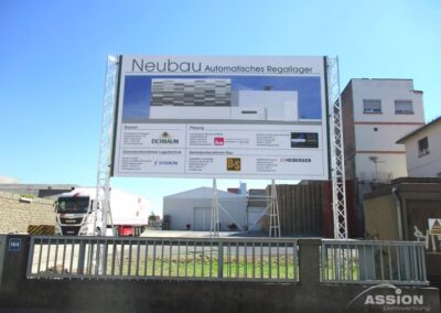 Großes Bauschild vor einem Industriegebiet mit Informationen über ein neues automatisches Lager, einschließlich Projektpartnern und einer Abbildung des Gebäudes.