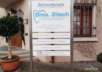 Ein Praxisschild mit der Aufschrift "Dres. Zitsch" und Mitarbeiter, Sprechzeiten, Kontaktinformationen und Stiftungsdetails, steht vor einem Gebäude.