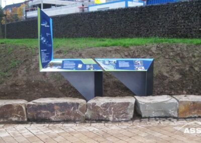 Informationsschild mit Text und Bildern auf Metallträgern vor einer grasbewachsenen Böschung und Steinsitzblöcken auf einer gepflasterten Fläche.