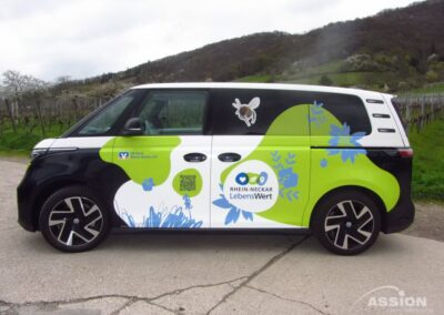 Ein Lieferwagen mit grüner, schwarzer und weißer Blumengrafik und dem Schriftzug "Rhein-Neckar LebensWert" ist im Freien in der Nähe von Bäumen und Hügeln geparkt.