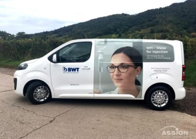Ein weißer Lieferwagen der Firma BWT mit einer Werbung, die eine Frau mit Brille und Laborkittel zeigt und auf der Seite für "WFI - Water for Injection" wirbt.