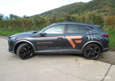 Ein grauer Geländewagen mit dem Schriftzug "Falkenhain Metall Design" ist auf einer Landstraße mit Grünflächen und Hügeln im Hintergrund geparkt.