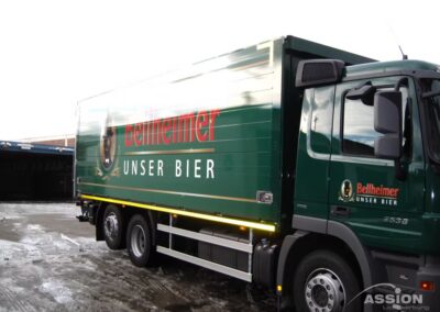 Ein grüner Lieferwagen mit dem Schriftzug "Bellheimer Unser Bier" parkt auf dem nassen Bürgersteig neben einem Gebäude.