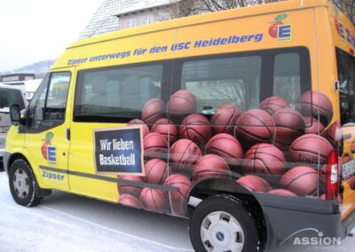 Ein gelber Lieferwagen mit Basketball-Grafiken und deutschem Text, der für die Unterstützung des USC Heidelberg und die Liebe zum Basketball wirbt, parkt auf einer verschneiten Straße.
