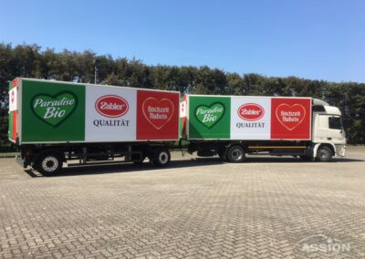 Zwei weiße Lastwagen mit großen grünen und roten Kartons mit den Logos "Paradiso Bio", "Zabler" und "Qualität" sind auf einem gepflasterten Parkplatz mit Bäumen im Hintergrund geparkt.