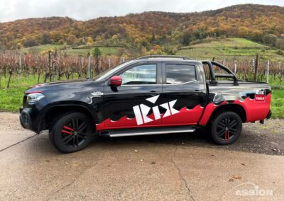 Ein schwarz-roter Pickup mit "Kix"-Schriftzug parkt im Herbst auf einer Straße vor Weinbergen und bewaldeten Hügeln.