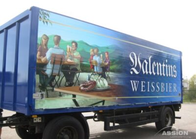 Blauer Lastwagen mit dem Schriftzug "Valentinus Weissbier" und einer Wandmalerei, die Menschen beim Biertrinken an Tischen im Freien zeigt, mit einem Tablett mit Würstchen und Gurken im Vordergrund.