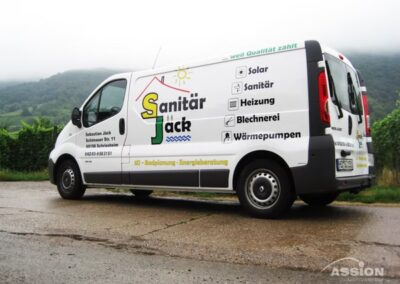 Ein weißer Lieferwagen mit dem Schriftzug "Sanitär Jack" und den Kontaktdaten ist auf einer Landstraße geparkt, mit grünen Feldern und Hügeln im Hintergrund.