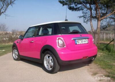 Ein rosafarbener Mini Cooper mit weißem Dach ist auf einer gepflasterten Straße neben Bäumen und einem Drahtzaun geparkt, von der hinteren linken Seite aus gesehen.