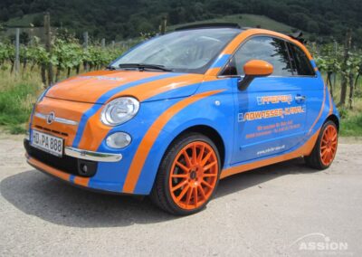 Ein blau-orangefarbener Fiat 500 mit orangefarbenen Rädern und einem Werbetext auf der Tür ist in der Nähe eines Weinbergs geparkt.