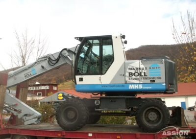 Ein kompakter Mobilbagger mit der Aufschrift "Markus Böll Bauunternehmung" wird auf einen roten Tieflader geladen, im Hintergrund sind Gebäude und Bäume zu sehen.