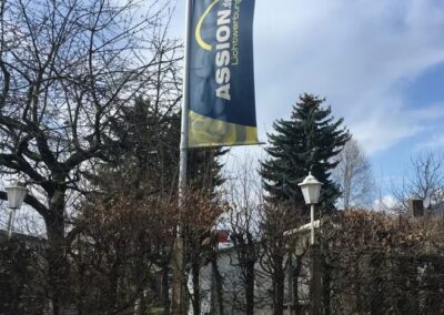 Ein großes Banner mit der Aufschrift ASSION" steht in der Nähe von blattlosen Bäumen und einem Drahtzaun, im Hintergrund sind Häuser und eine Straßenlaterne zu sehen, der Himmel ist teilweise bewölkt.