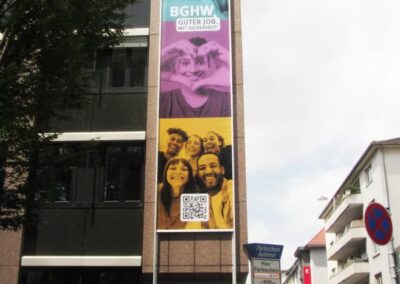 Ein großes Banner an einer Gebäudefassade zeigt das BGHW-Logo, den Slogan, den QR-Code und Fotos von lächelnden Menschen, darunter Autos und Fußgänger auf der Straße.