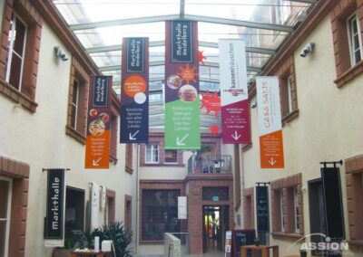 Innenkorridor mit hängenden Schildern mit Wegbeschreibung und Essensangeboten in deutscher Sprache, unter einer Glasdecke, mit Fenstern und Türen an den Wänden.