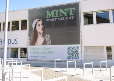 Ein großes Außenplakat an einem Gebäude zeigt eine junge Frau mit Mütze, die nach oben blickt, mit einem Text, der für Berufe und Ausbildung in den MINT-Fächern (MINT) auf Deutsch wirbt.