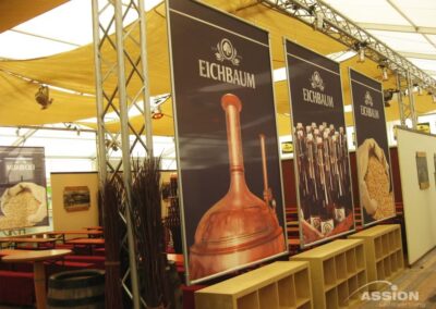 Innenraum eines Bierzeltes mit großen Bannern der Eichbaum-Brauerei, Holztischen, -bänken und -regalen unter einem gelben Vordach.