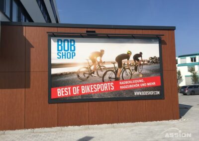 Große Außenwerbung für "Bob Shop" zeigt Radfahrer, die bei Sonnenuntergang auf einer Straße fahren, und wirbt für Radsportbekleidung und -zubehör in deutscher Sprache.