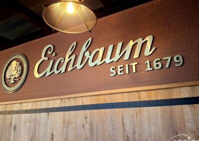 Auf einem Schild an einer Holzwand steht "Eichbaum SEIT 1679" in eleganten Einzelbuchstaben mit einem Baumlogo, beleuchtet von einer Deckenlampe.